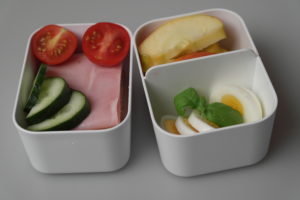Matboks med egg, eplebåter og brødskiver med skinke pyntet med tomat og agurk